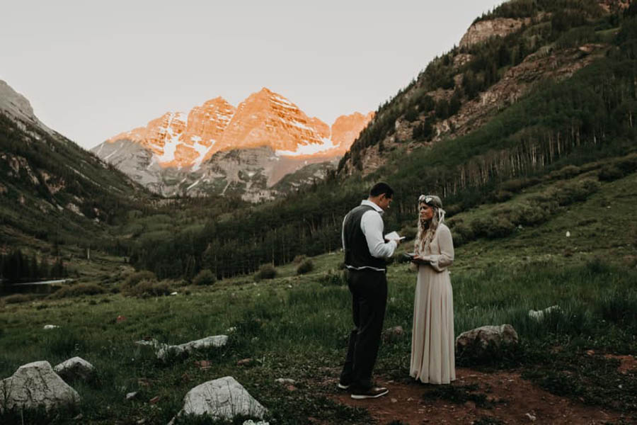 A wedding couple is reading their vows to each other and standing in a valley with the mountains and a forest behind them at the Maroon Bells.