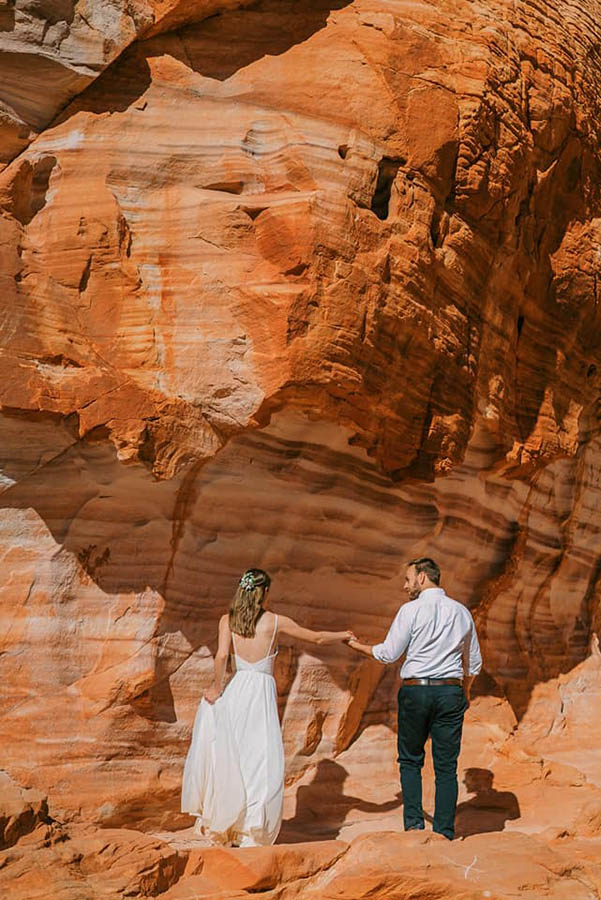 A wedding couple is holding hands and facing a orange mountain at the Valley of Fire State Park.