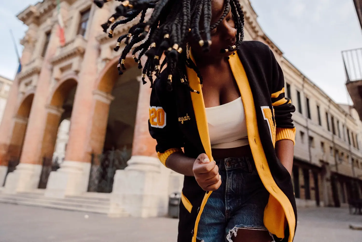 Woman in a varsity jacket with braids in motion: A dynamic shot of a woman wearing a black and yellow varsity jacket over a white top and denim shorts. Her beaded braids swing as she moves, set against the backdrop of an old stone building with large arches, creating a sense of energy and style.