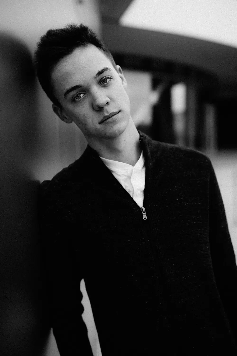 Black-and-white portrait of a young man: A black-and-white close-up of a young man leaning against a wall, wearing a dark zip-up sweater. He gazes directly at the camera with a calm, introspective expression, set against a blurred urban background.