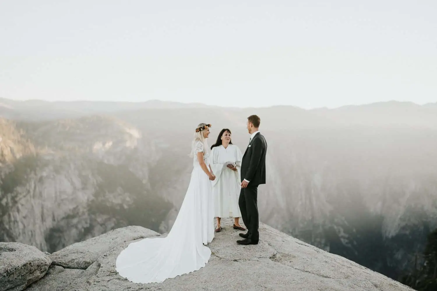 Bride and groom exchanging vows on a cliff during a Yosemite elopement ceremony, with an officiant and misty mountains in the background.